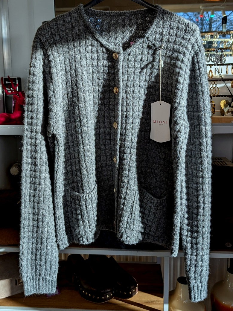 Die graue Cardigan-Strickjacke im Chanel-Stil mit ihren markanten Quadrate und edlen Goldknöpfen vereint zeitlose Eleganz mit modernem Komfort!